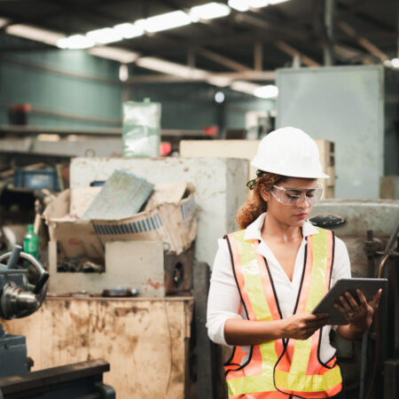 Female industrial engineer wearing a white helmet while standing in a heavy industrial factory behind she looking of working at industrial machinery and check security system setup in factory.